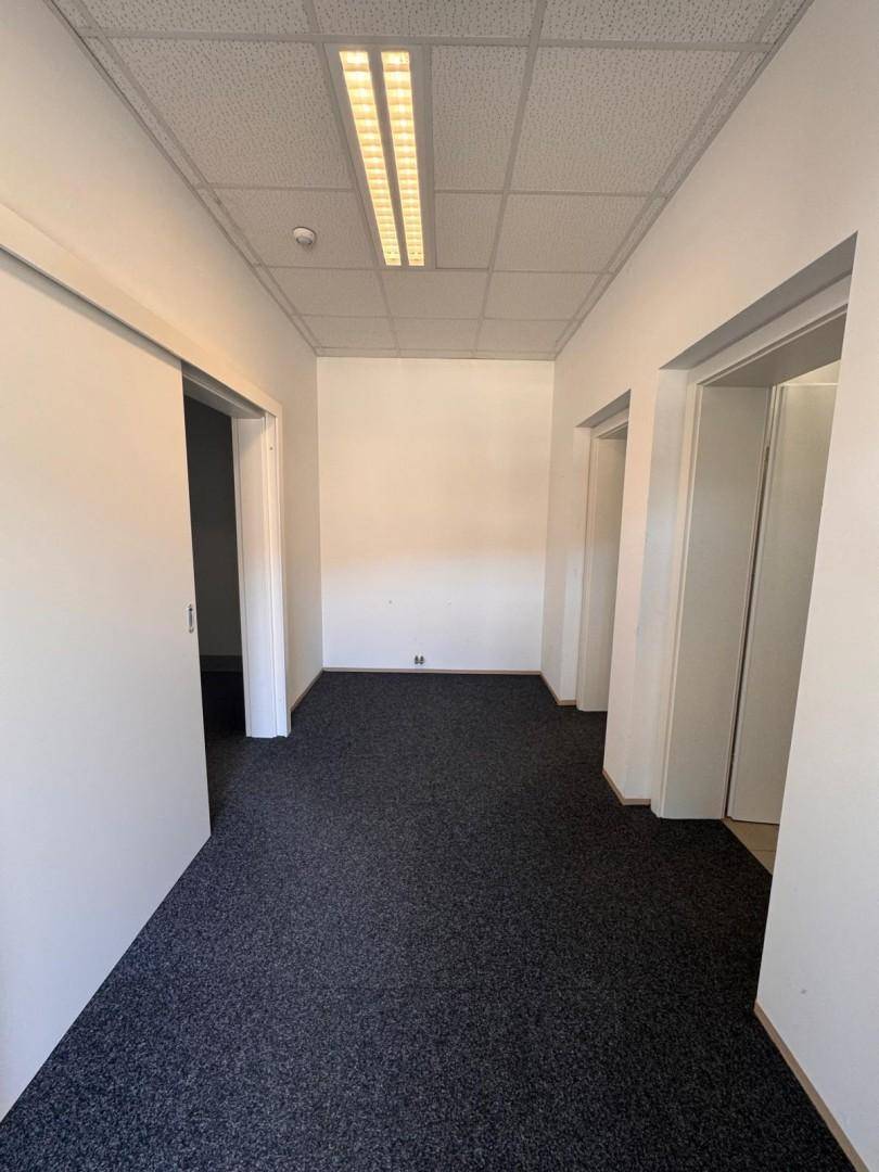 Langer Flur mit Teppichboden, weißen Wänden und Deckenleuchten in einem Bürogebäude.