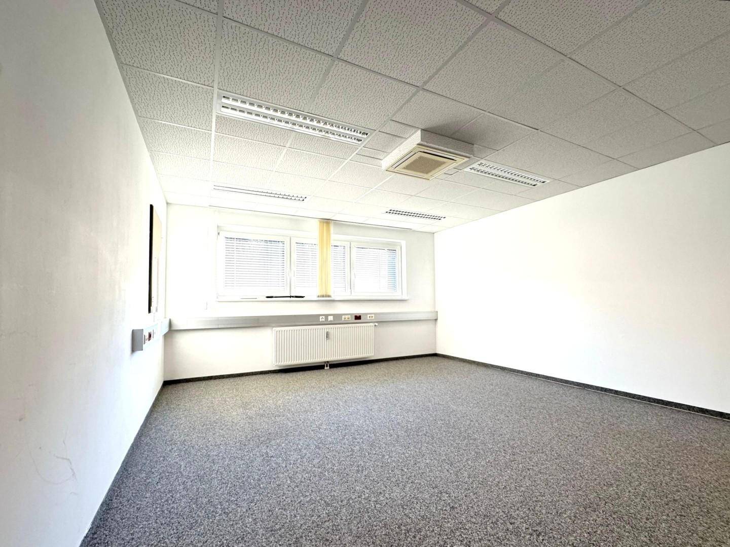 Leeres Büro mit grauem Teppichboden, weißen Wänden und Fenstern, die für natürliches Licht sorgen, sowie einer Klimaanlage an der Decke.