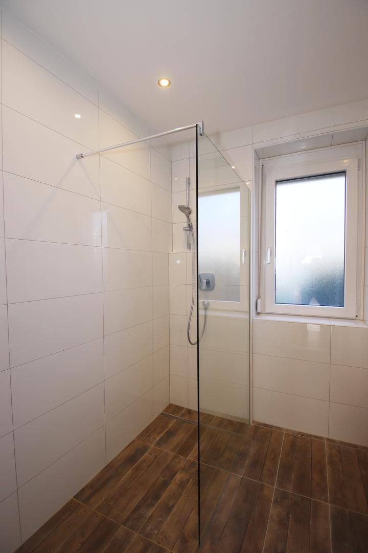 Duschbereich im Badezimmer mit Glasabtrennung, weißen Fliesen und Fenster für natürliche Belüftung.