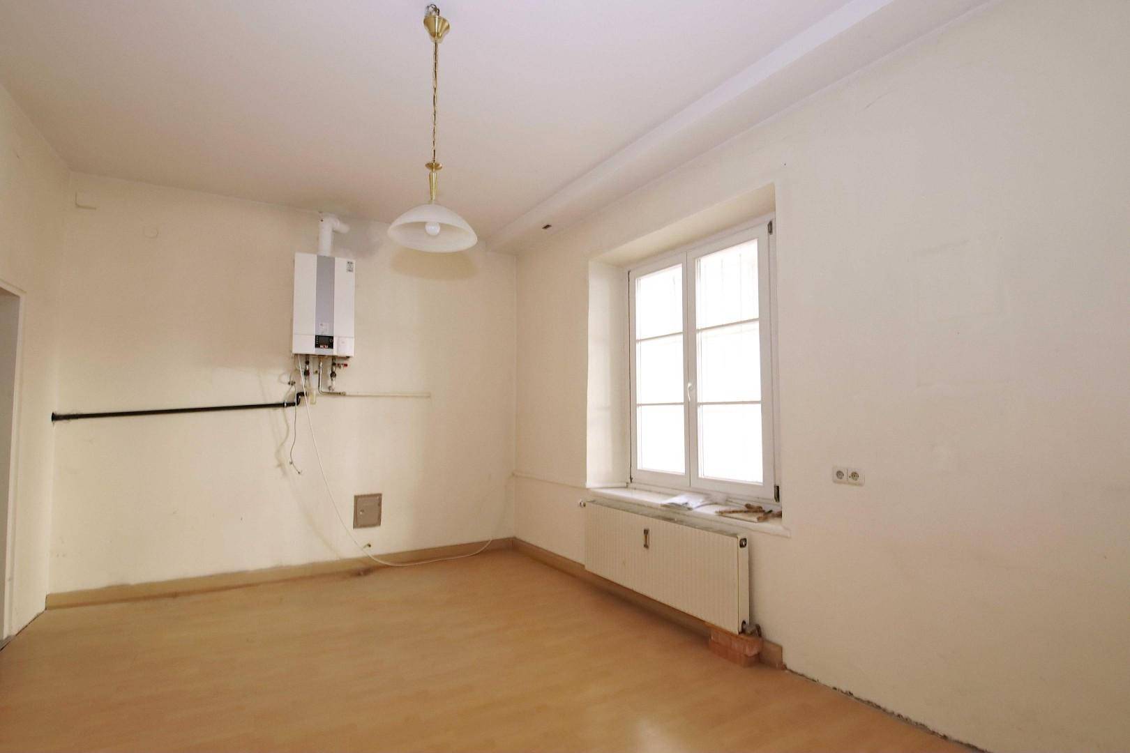 Leerer Raum mit hellem Laminatboden, einem Fenster, Heizkörper und einem an der Wand montierten Warmwasserbereiter.