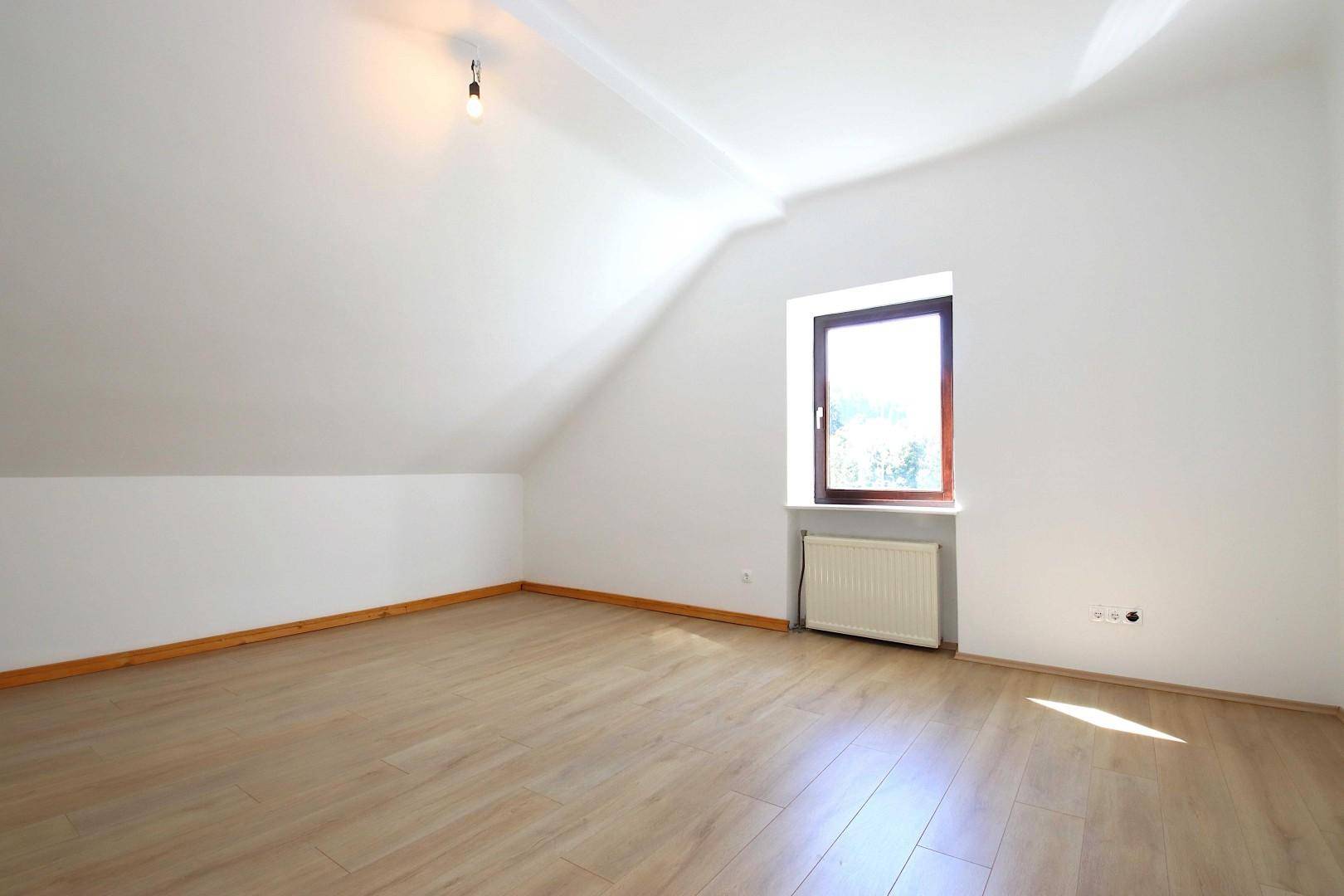 Leerer Raum mit Laminatboden, Dachschräge und einem Fenster, das viel Tageslicht hereinlässt.