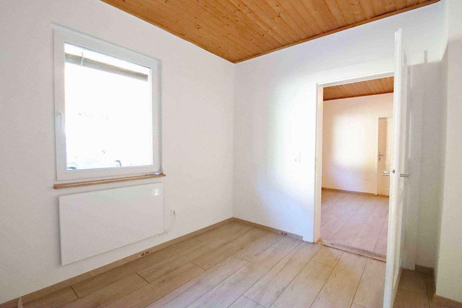 Helles Zimmer mit Holzboden und Fenster, das einen Zugang zu einem weiteren Raum bietet.