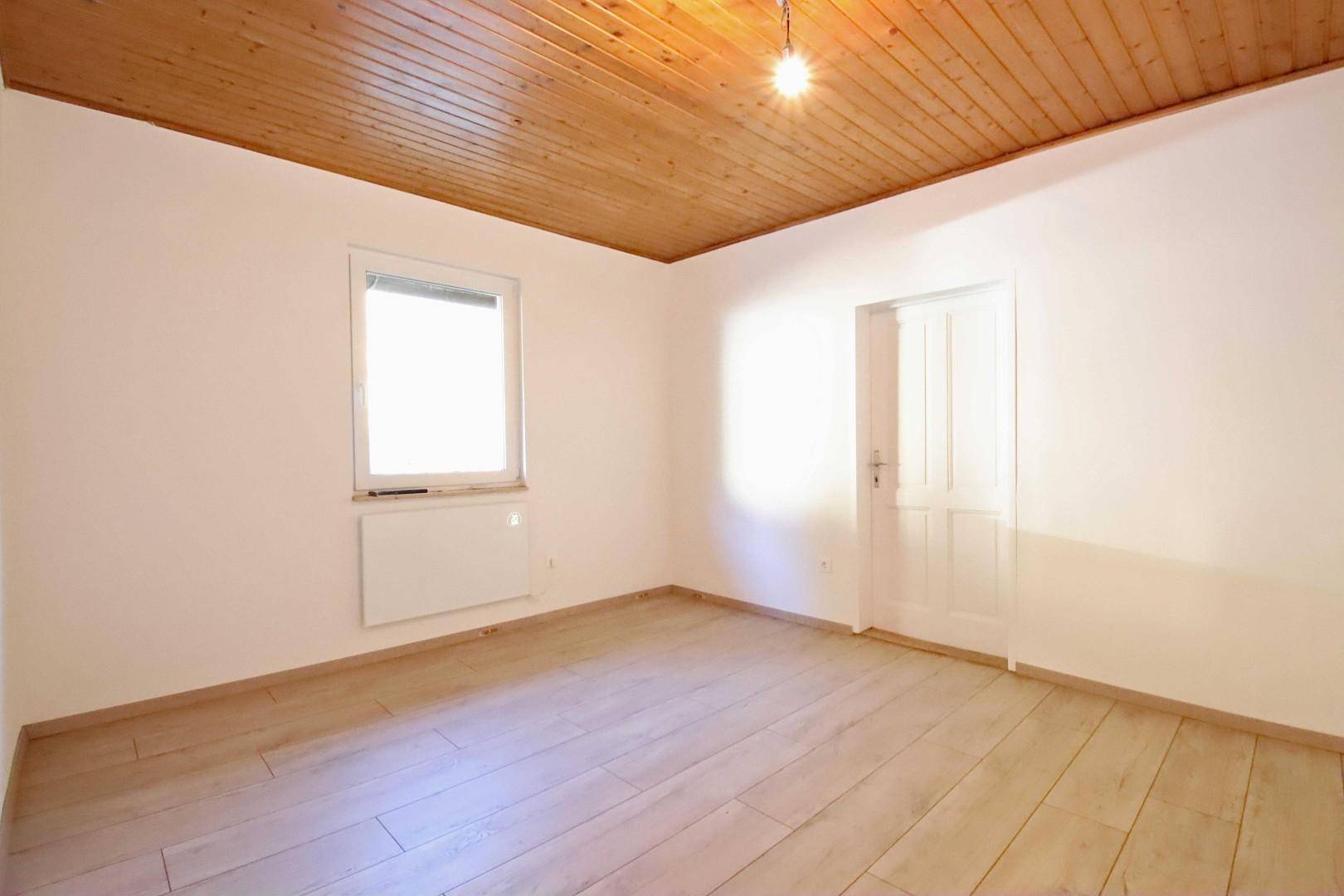 Leeres Zimmer mit hellem Holzboden, Fenster und Heizkörper unter einer Holzpaneeldecke.
