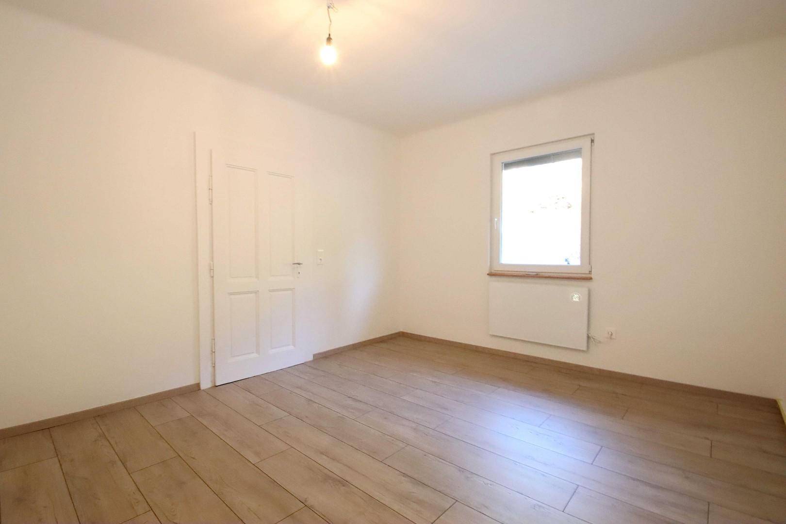 Geräumiges Zimmer mit hellem Holzboden, weiß gestrichenen Wänden und einem Fenster.