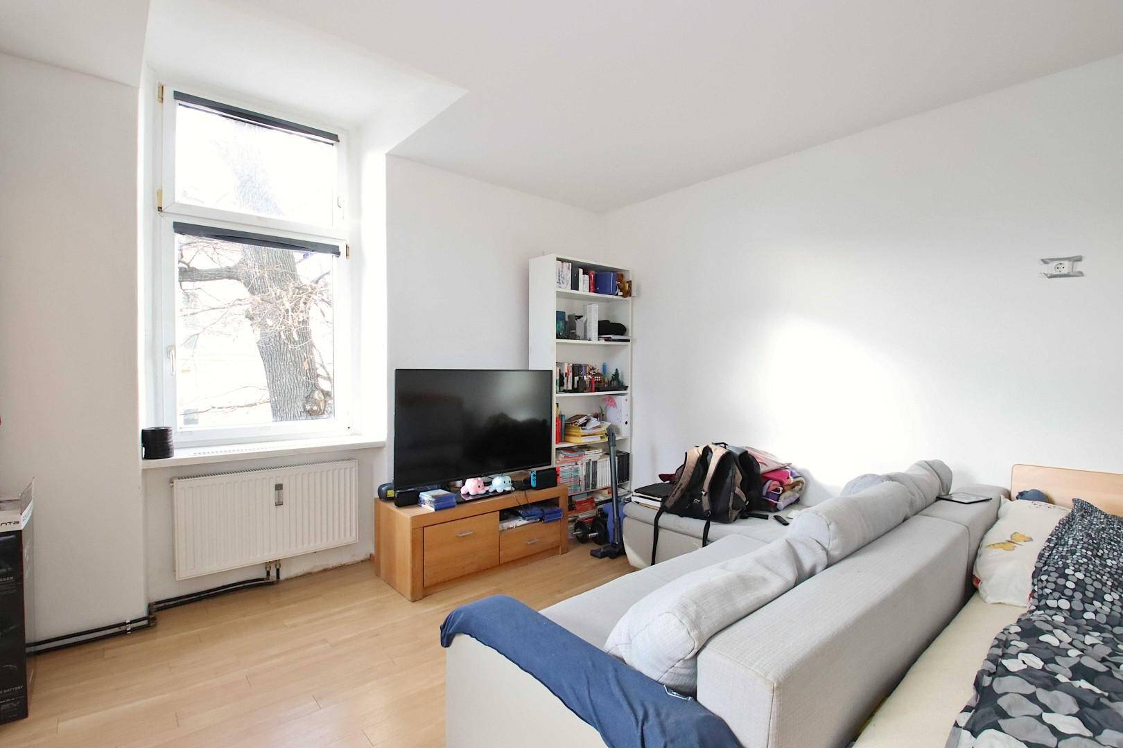 Gemütliches Wohnzimmer mit Sofa, Fernseher und großem Fenster, das viel Tageslicht hereinlässt.