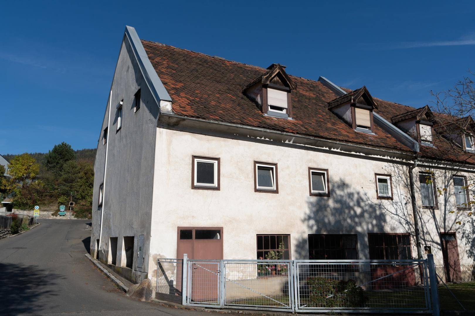 Älteres Gebäude mit Satteldach und kleinen Fenstern an einer ländlichen Straße im Sonnenlicht.