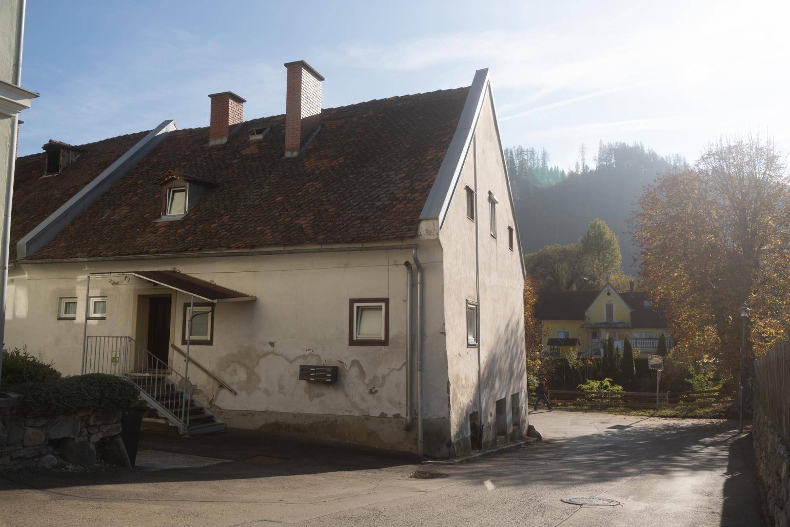 Älteres Gebäude mit Satteldach, zwei Schornsteinen und einer Außentreppe an einer Straße.