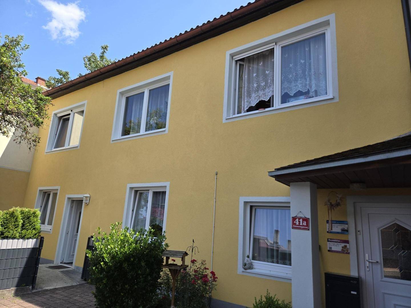 Seitenansicht der gelben Hausfassade mit gepflegtem Vorgarten und Eingangsbereich an einem sonnigen Tag.