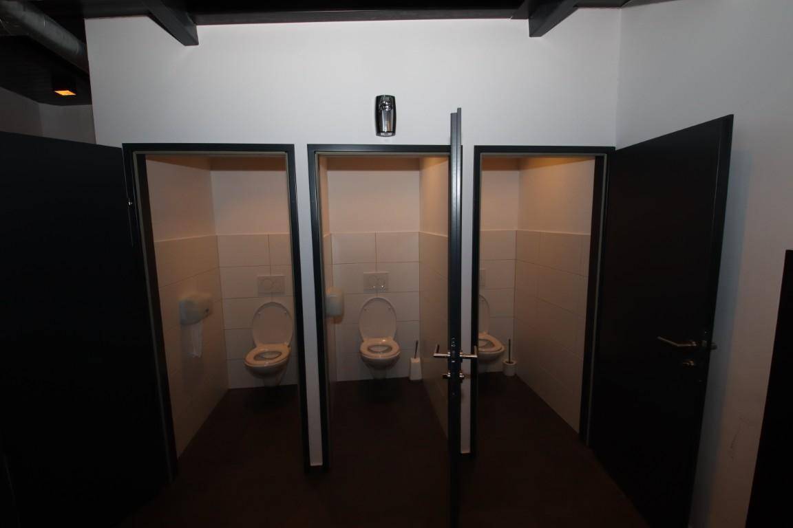 Damen-WC mit drei Toilettenkabinen, weißen Fliesen und dunklen Türen.