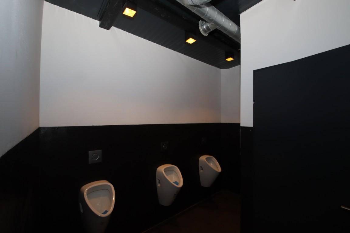 Herren-WC mit drei Urinalen, schwarzen und weißen Wänden und sichtbaren Rohren an der Decke.