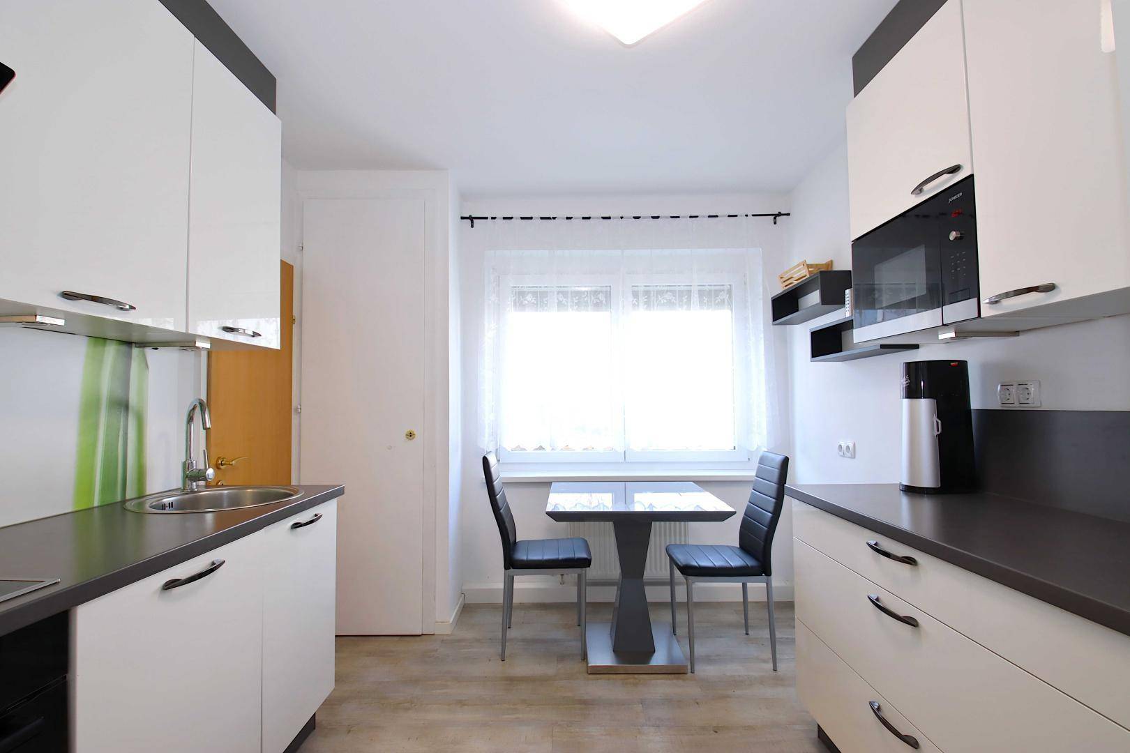 Moderne Küche mit weißen Schränken, dunkler Arbeitsplatte und kleinem Essbereich am Fenster.