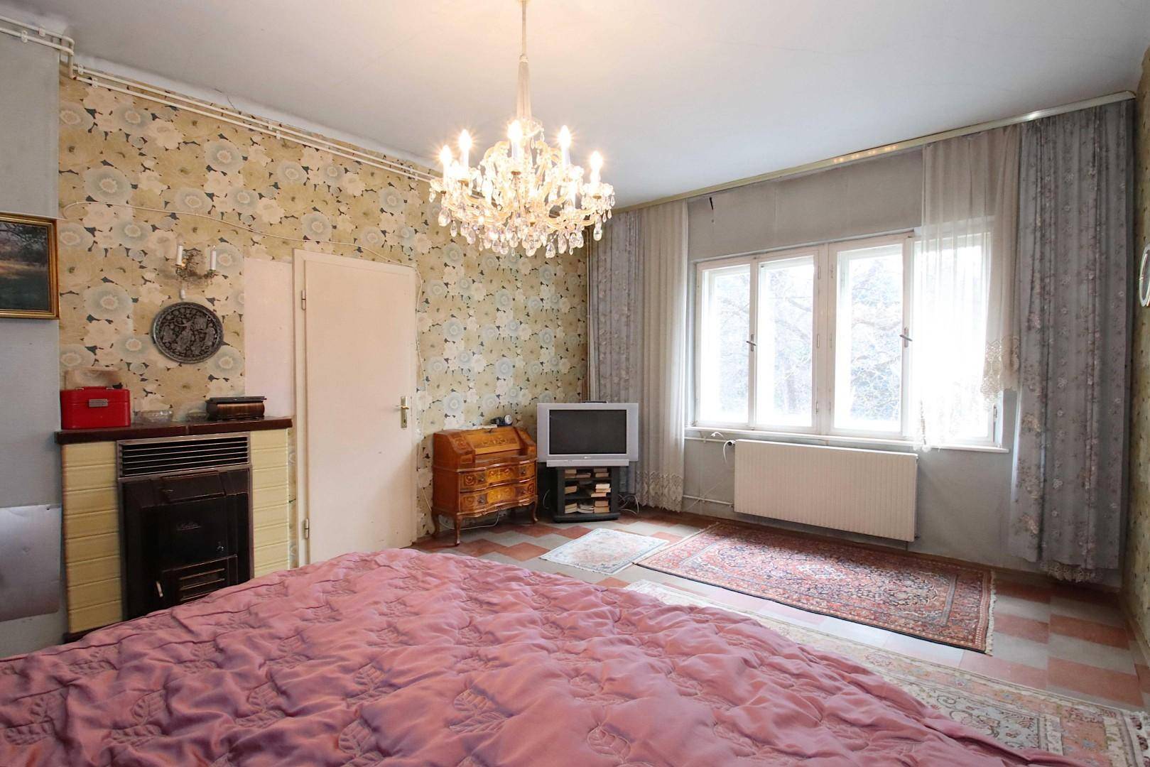 Schlafzimmer mit floraler Tapete, Kachelofen, Kronleuchter und Blick ins Grüne.