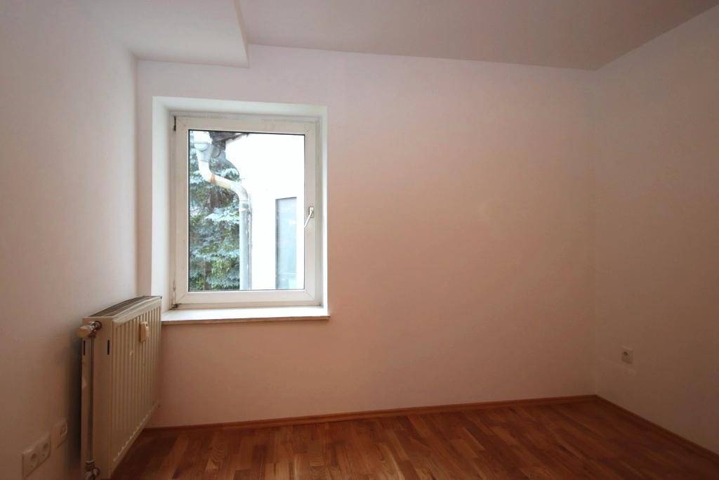 Unmöblierter Raum mit Holzparkett und Fenster, das Tageslicht hereinlässt und einen Blick nach draußen bietet.