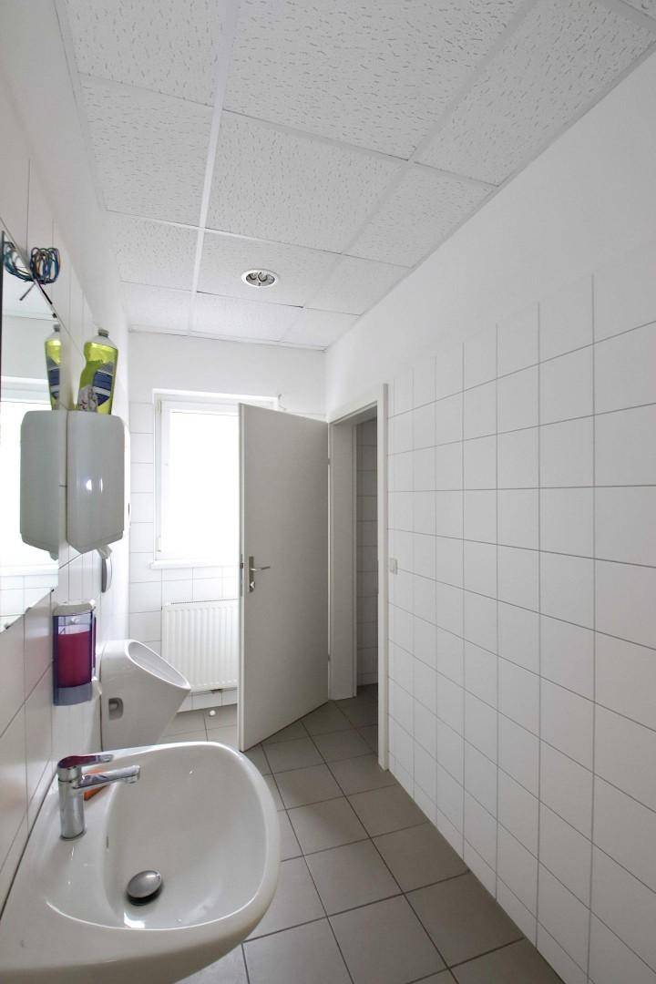 Helles Badezimmer mit weißen Fliesen, Waschbecken und Urinal, gut beleuchtet durch ein Fenster.