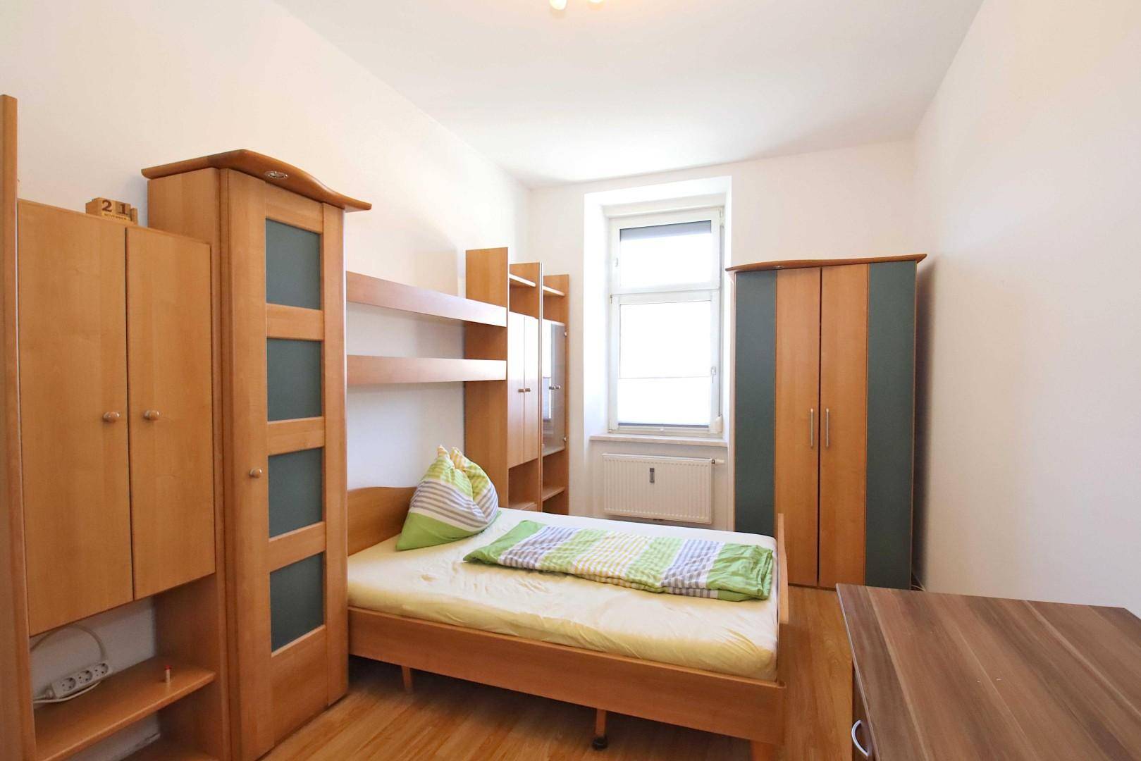 Schlafzimmer mit Einzelbett, Holzmöbeln und Laminatboden, hell durch ein Fenster.