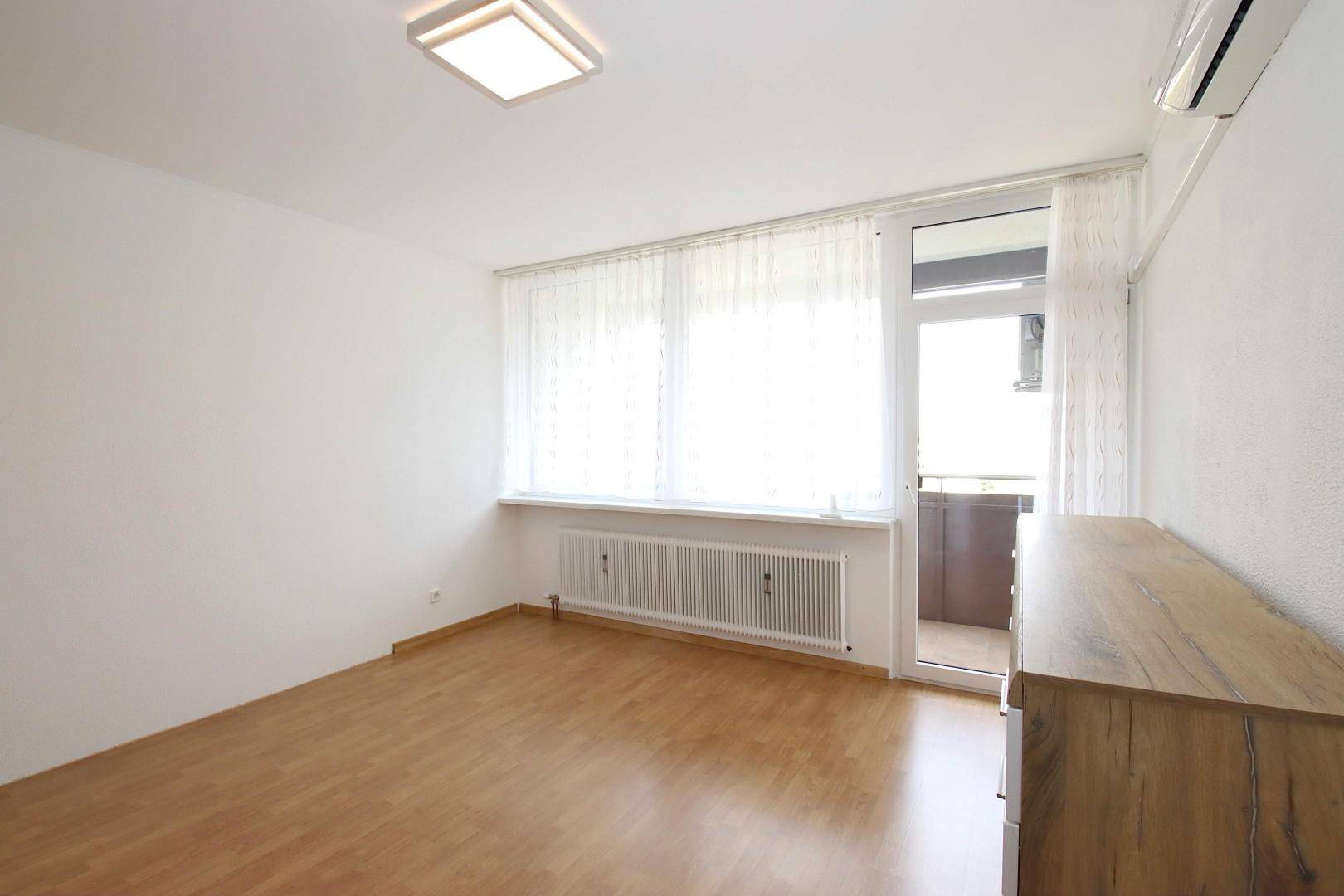 Helles Zimmer mit Laminatboden, Fenster und Zugang zu einem Balkon, ausgestattet mit Klimaanlage.