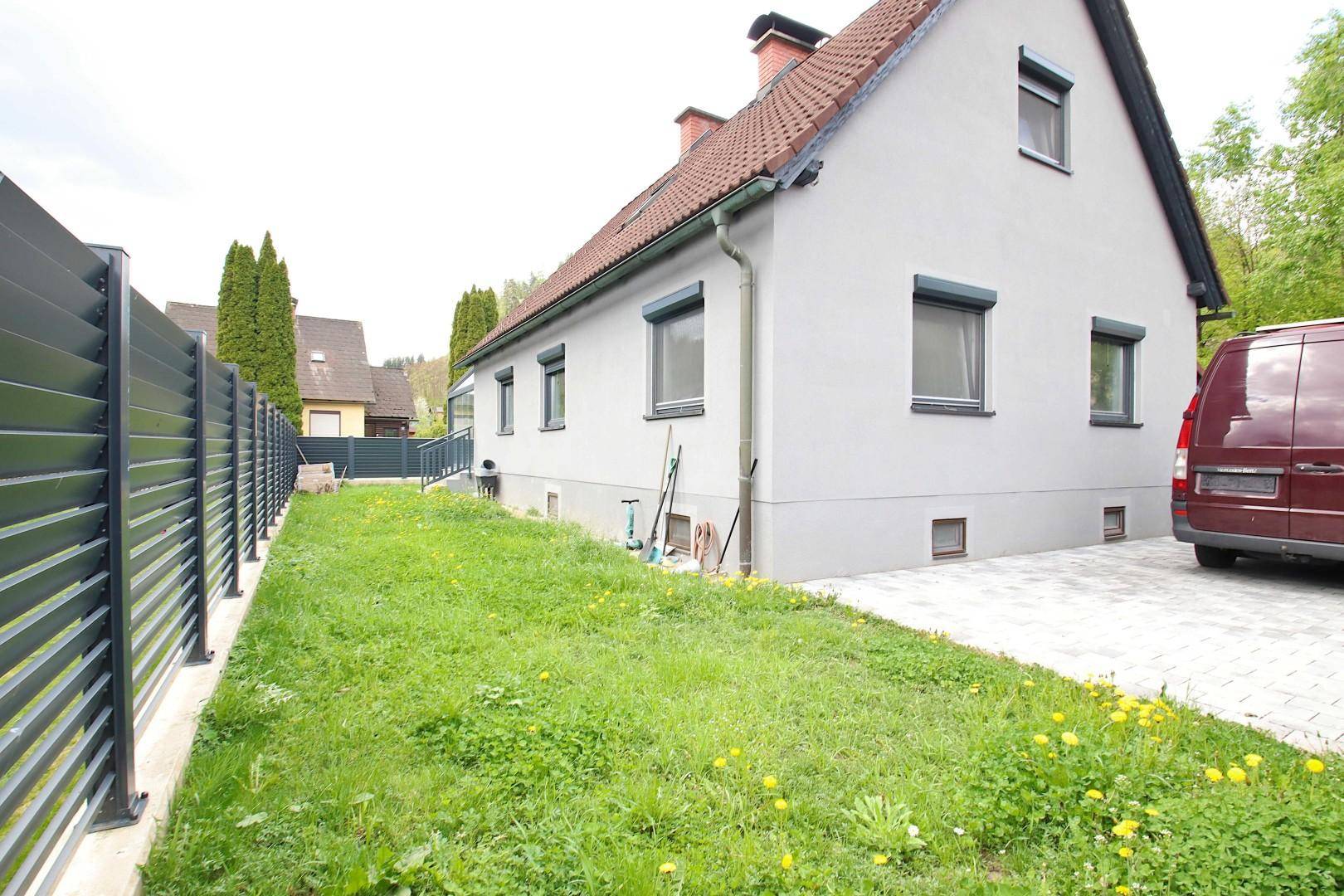 Seitenansicht des Hauses mit Einfahrt, Zaun und grünem Rasenbereich.
