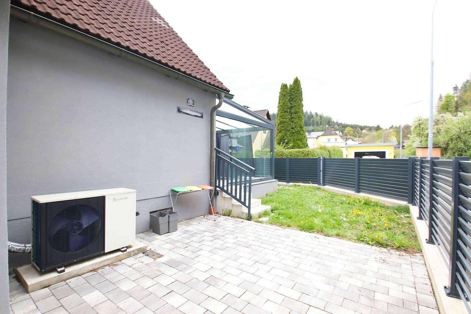 Seitenansicht des Hauses mit Wärmepumpe, gepflastertem Bereich und kleinem Rasenstück.
