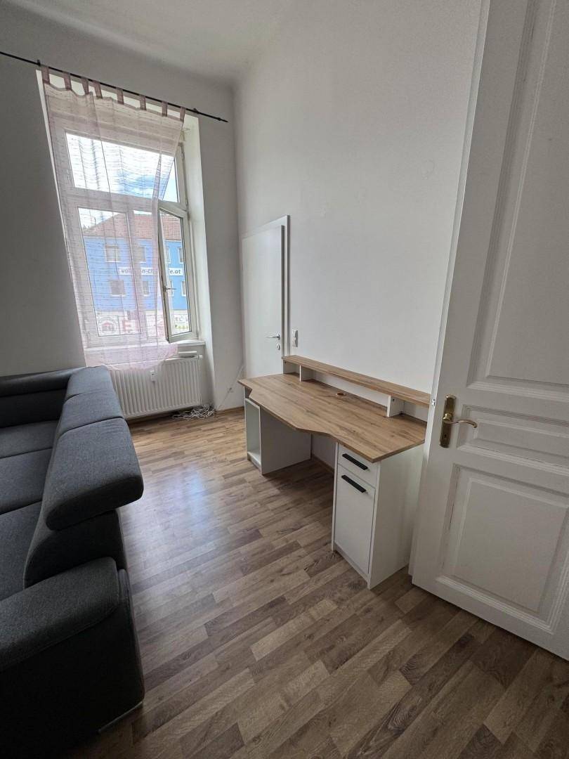 Helles Wohnzimmer mit Sofa, Schreibtisch, Laminatboden und großem Fenster.