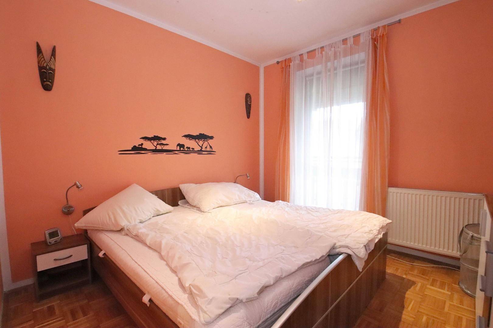 Schlafzimmer mit orangefarbener Akzentwand, Doppelbett und Parkettboden.