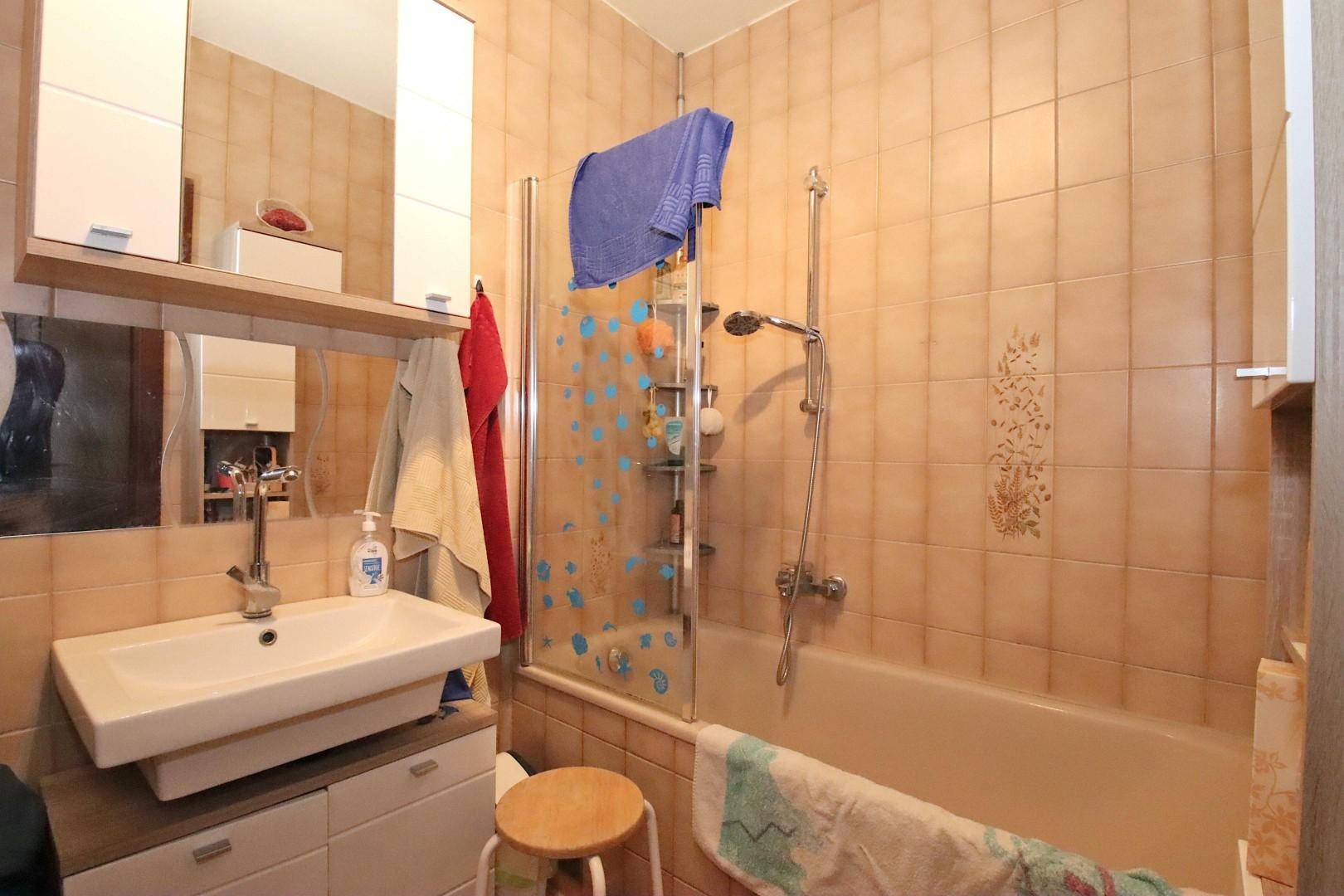 Badezimmer mit beigefarbenen Fliesen, Badewanne mit Duschfunktion und Einzelwaschbecken.