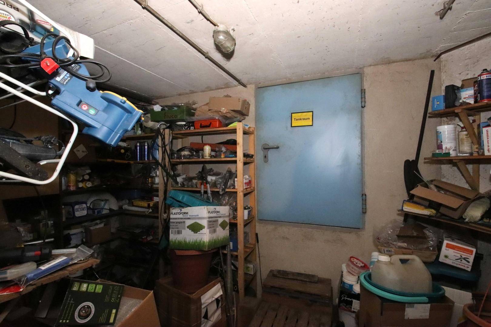 Überfüllter Abstellraum im Keller mit Regalen voller Gegenstände und einer Tür zum Tankraum.