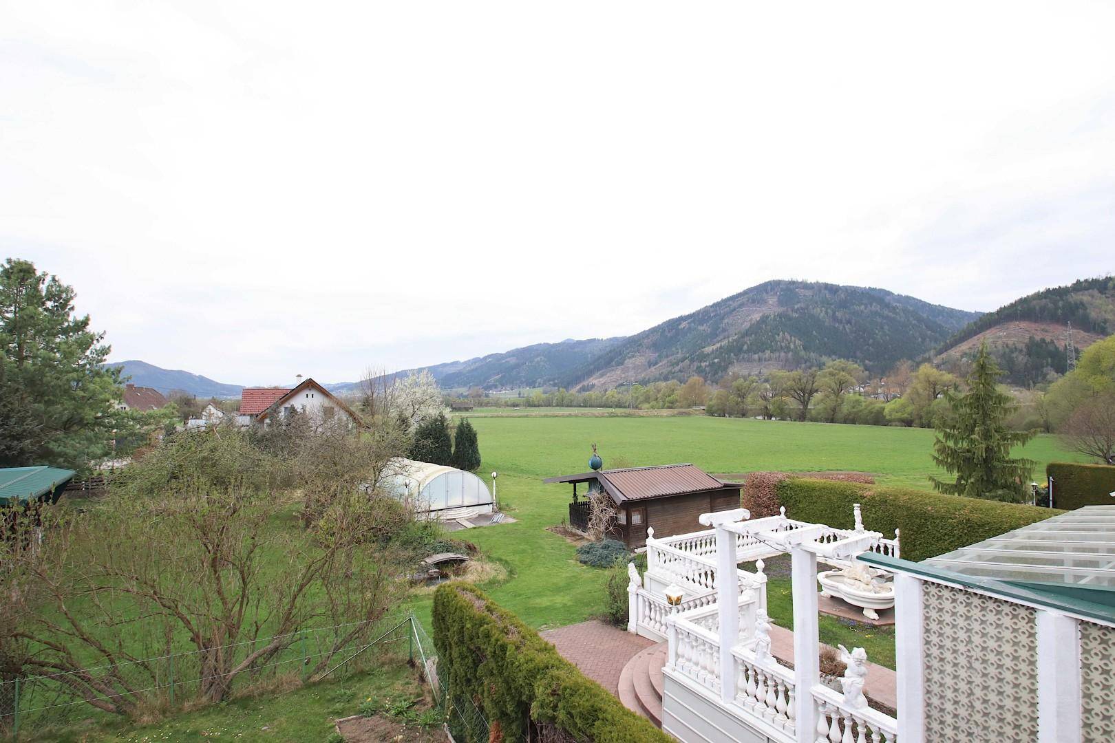 Panoramablick auf einen weitläufigen Garten, grüne Felder und die umliegende Berglandschaft.