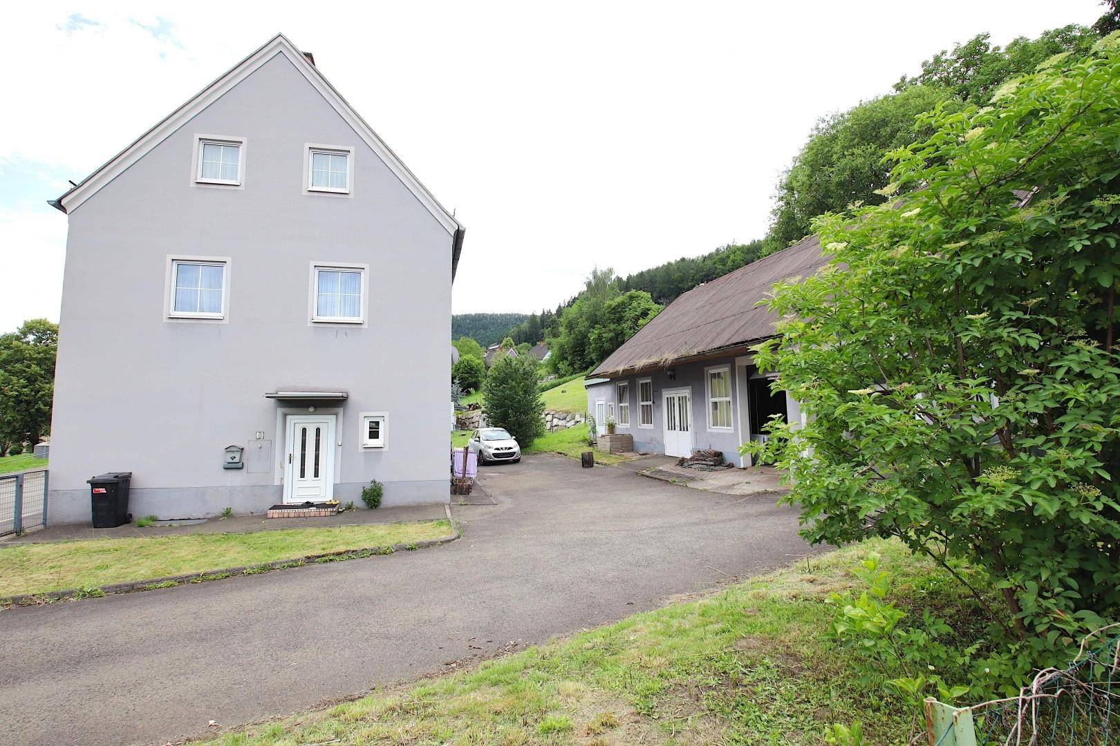 Ansicht des grauen Wohnhauses und des Nebengebäudes mit Zufahrt und grüner Umgebung.