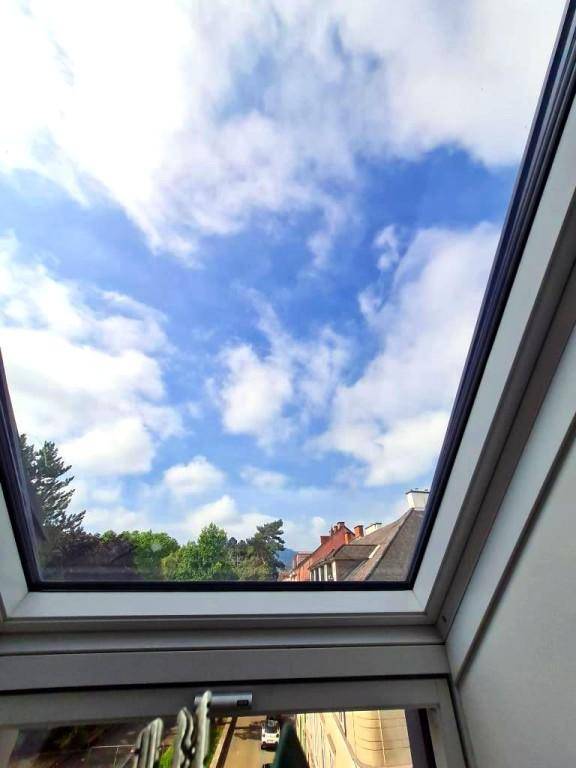 Blauer Himmel mit Wolken und Blick über Dächer und Bäume aus einem Dachfenster.