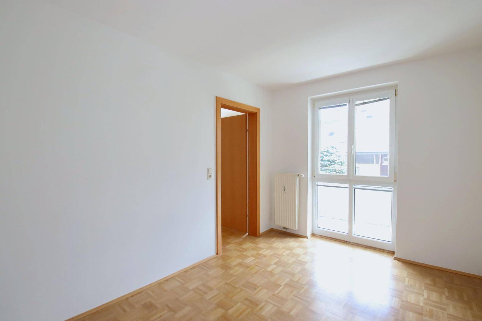 Helles Zimmer mit Parkettboden und großem Fenster, das viel Tageslicht hereinlässt.
