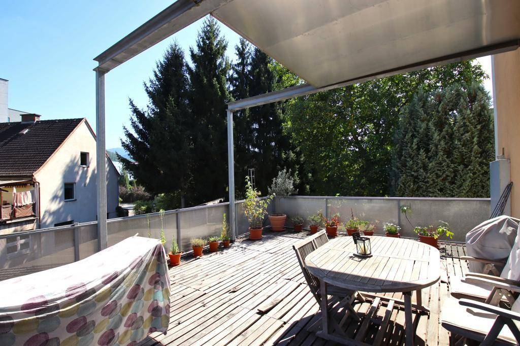 Geräumige Holzterrasse mit Esstisch, Stühlen und vielen Topfpflanzen unter einer schützenden Überdachung.