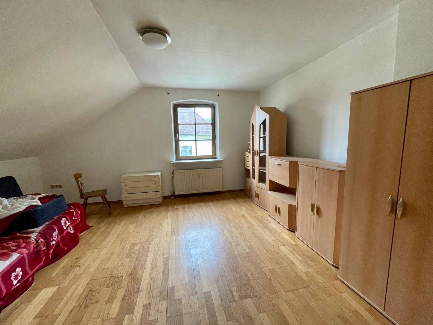 Helles Dachzimmer mit Holzfußboden, schrägen Wänden und einfacher Möblierung.