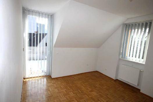 Dachzimmer mit schrägen Decken, Parkettboden und einem großen Fenster mit Jalousien.