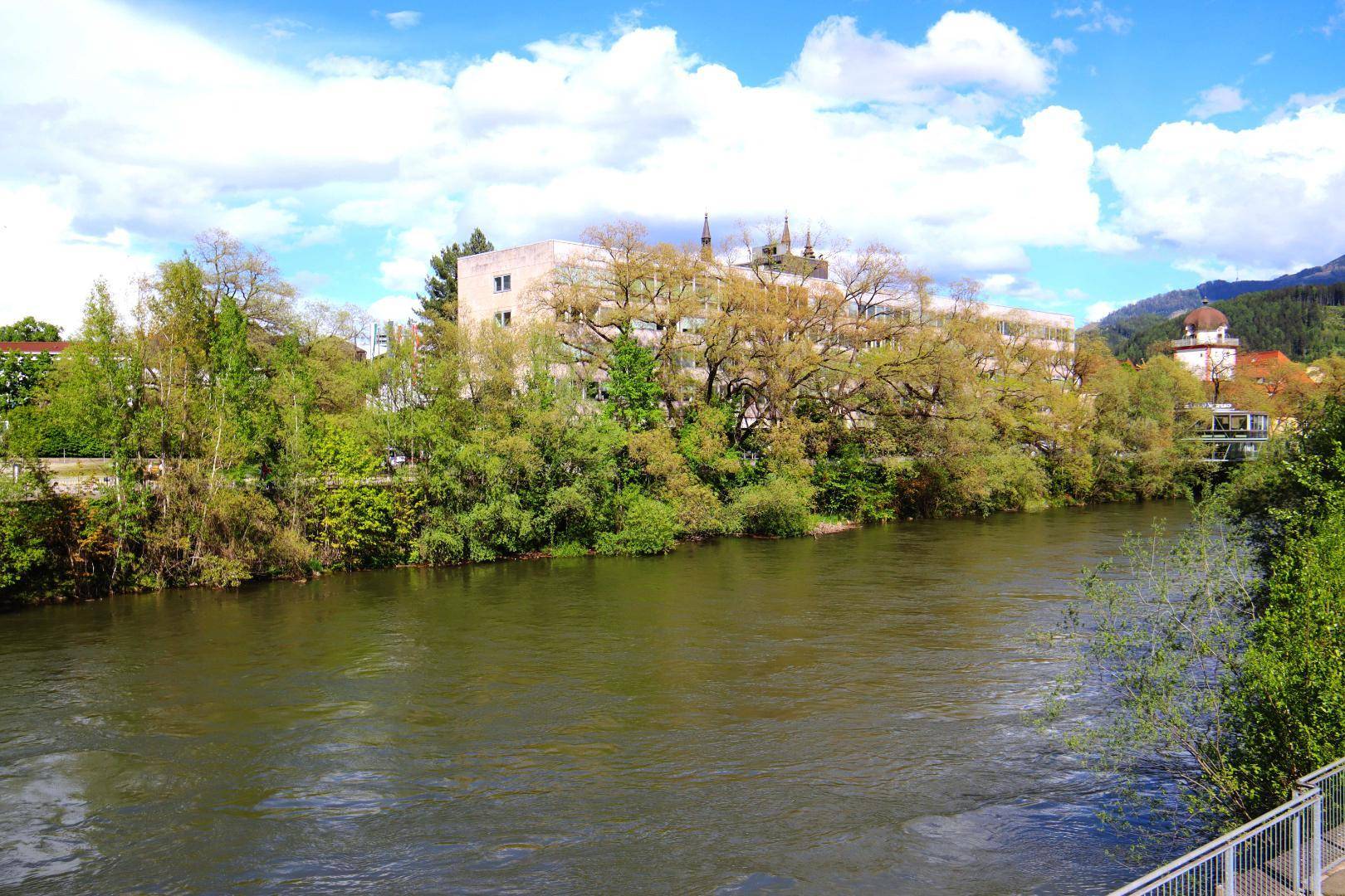 Breiter Fluss mit grünen Ufern, umgeben von Bäumen, Gebäuden und Bergen im Hintergrund.