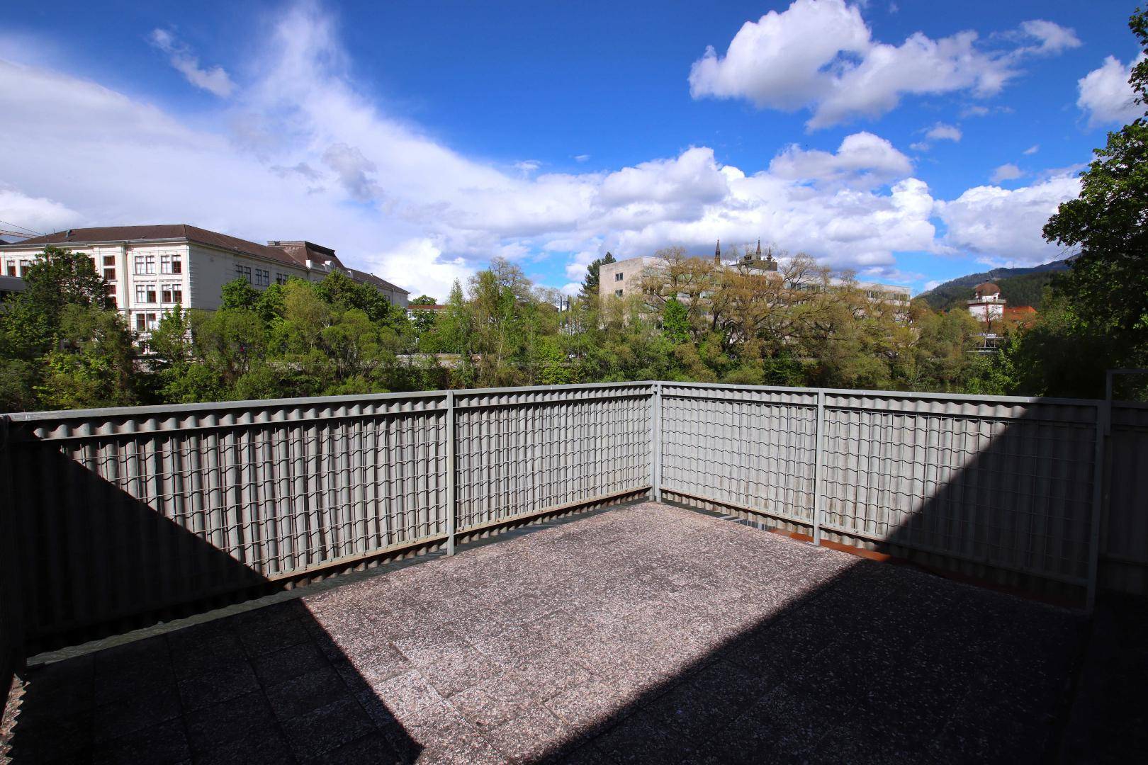 Leere Terrasse mit grauen Fliesen und Metallgeländer, bietet Blick auf Gebäude und Bäume.