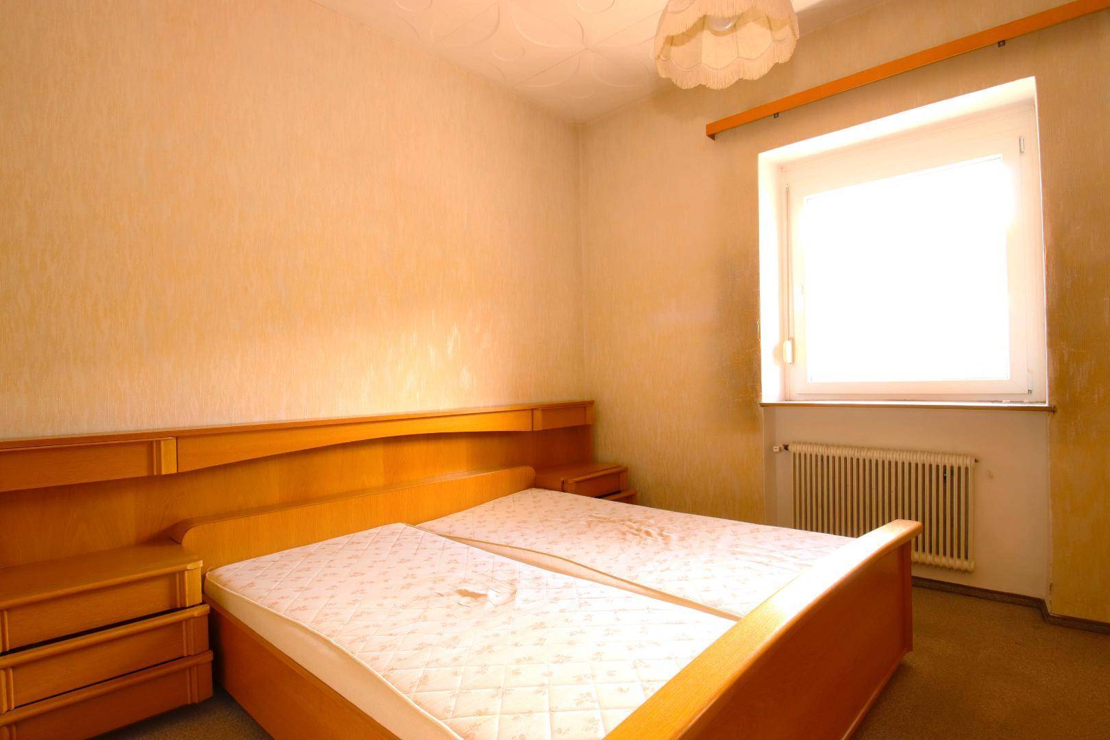 Schlafzimmer mit gelber Tapete, Doppelbett mit Holzrahmen und einem Fenster mit Heizkörper.