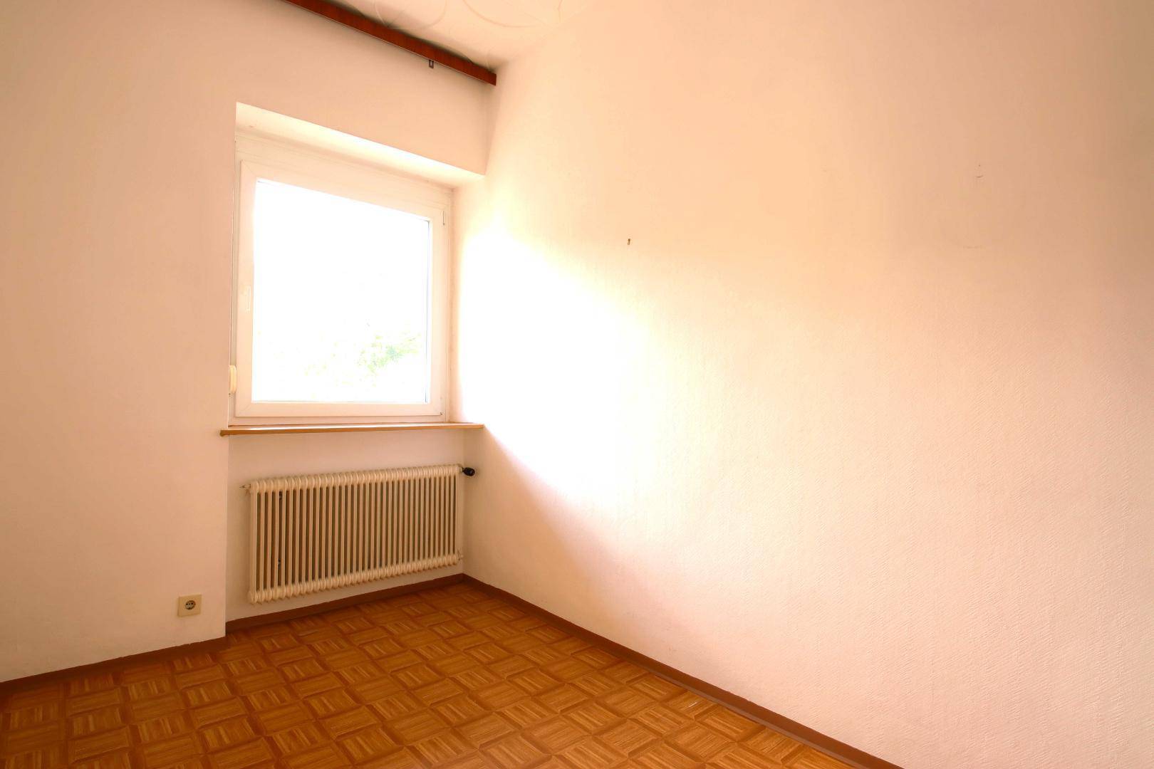 Leerer Raum mit weißen Wänden, Fenster und Heizkörper, ausgestattet mit Parkettboden.