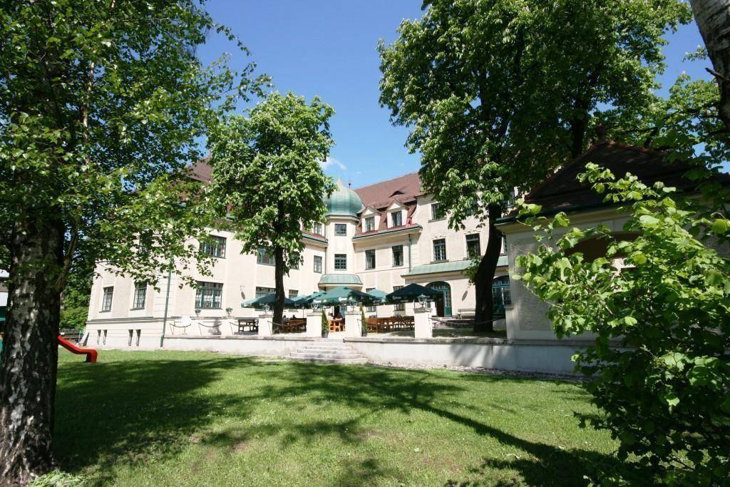 Historisches Gebäude mit weitläufigem Gartenbereich, schattenspendenden Bäumen und Außenterrasse mit Sitzgelegenheiten.