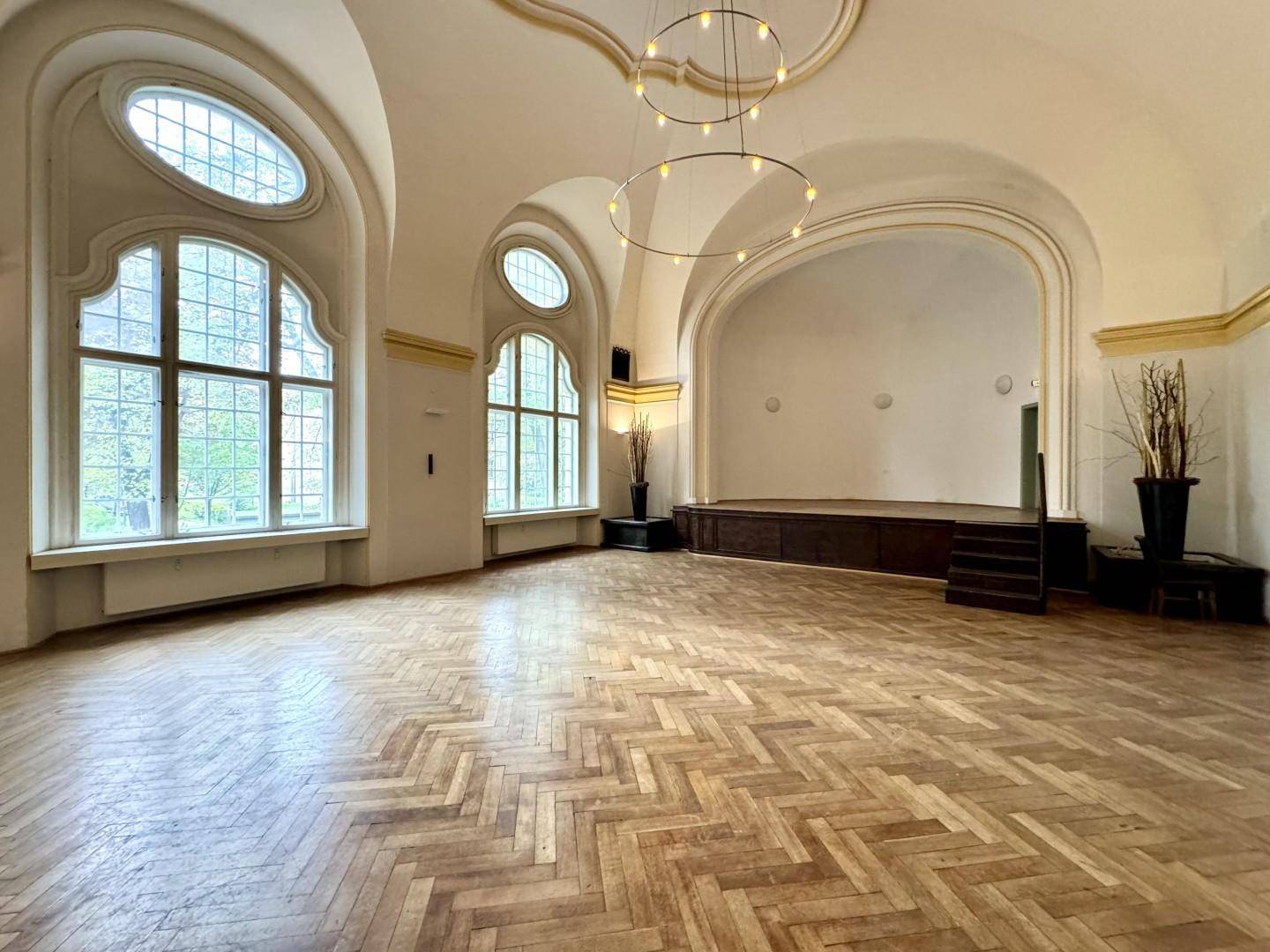 Geräumiger Veranstaltungssaal mit eleganter Fischgrätparkett und einer erhöhten Bühne, hell durch große Bogenfenster.