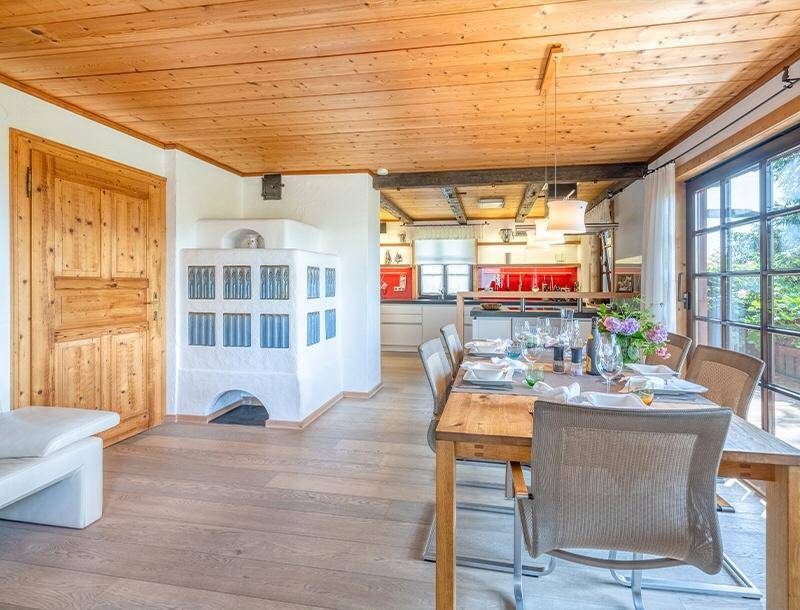 Heller Essbereich mit Holzbalkendecke, Kachelofen und offener Verbindung zur modernen Küche.