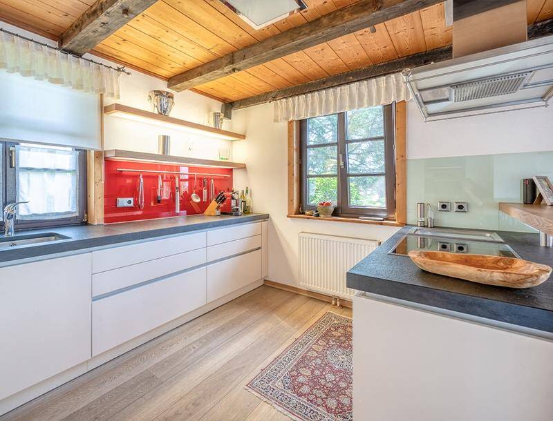 Moderne Einbauküche mit weißen Fronten, roter Glasrückwand und Holz-Arbeitsplatte unter Holzbalkendecke.