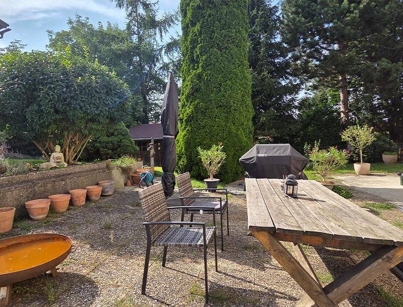 Gemütlicher Außenbereich mit Holztisch, Grillplatz und Feuerstelle, umgeben von viel Grün.