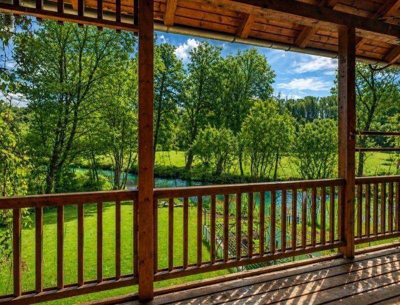 Blick von einem überdachten Holzbalkon auf einen malerischen Fluss und die umliegende grüne Naturlandschaft.