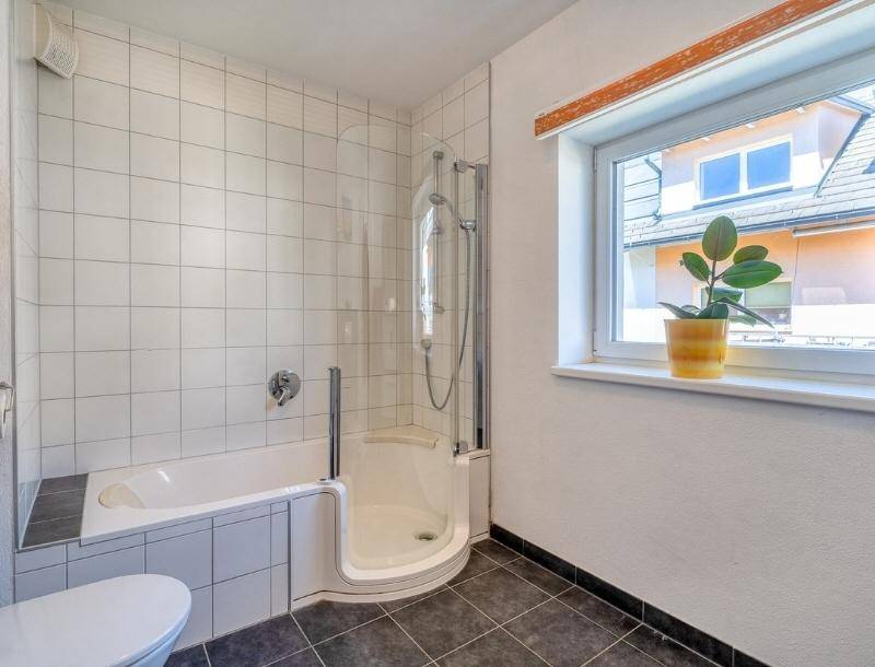 Badezimmer mit Badewanne, Dusche und Fenster, gefliest in Weiß und Anthrazit.
