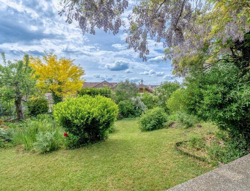 Gepflegter Garten mit Rasenflächen, blühenden Sträuchern und Bäumen unter bewölktem Himmel.