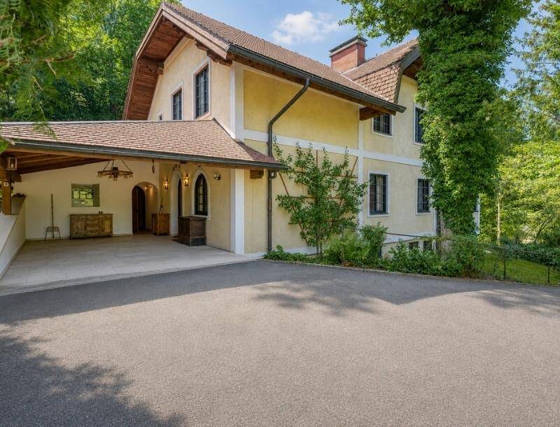 Seitenansicht des Hauses mit überdachtem Carport und breiter Zufahrt, umgeben von Bäumen.