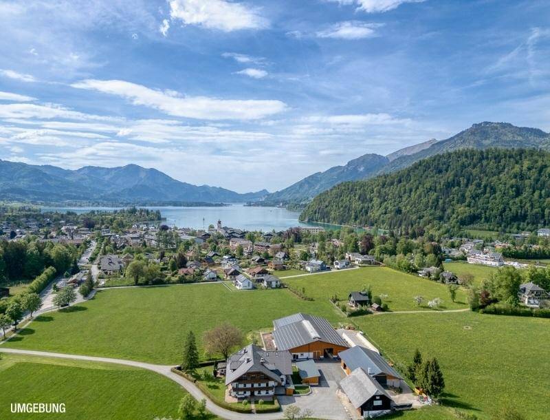 Weitläufige Luftaufnahme einer ländlichen Gegend mit Bauernhof, See und Bergen im Hintergrund.