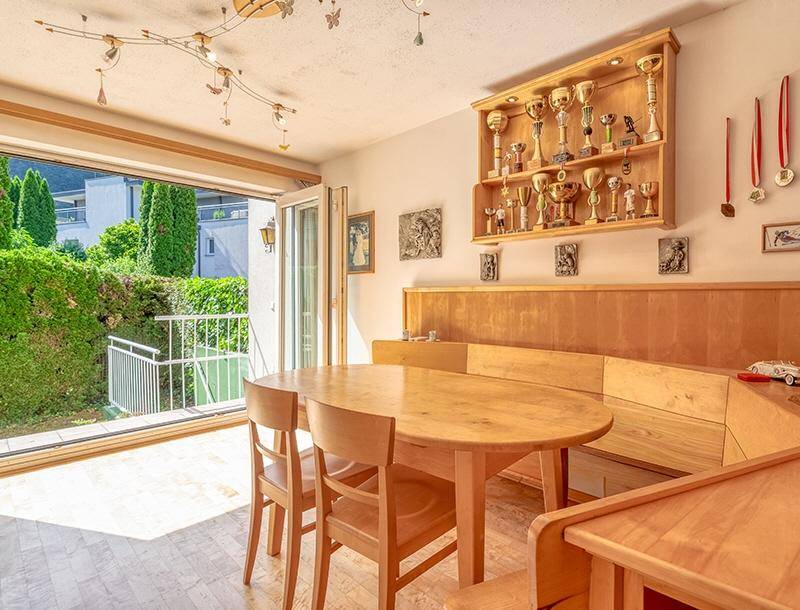 Gemütlicher Essbereich mit Holztisch, Eckbank und Zugang zu einem kleinen Balkon mit Heckenblick.
