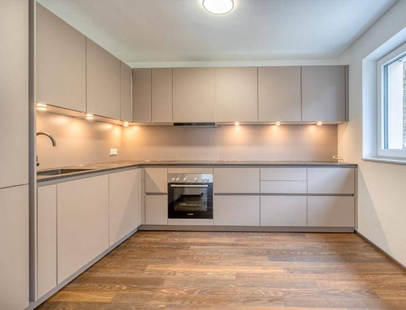 Moderne Einbauküche in Beige mit integriertem Backofen und Holzfußboden, hell durch ein Fenster.
