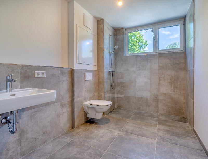 Modernes Badezimmer mit grauen Fliesen, wandhängendem WC, Waschbecken und einem Fenster für Tageslicht.